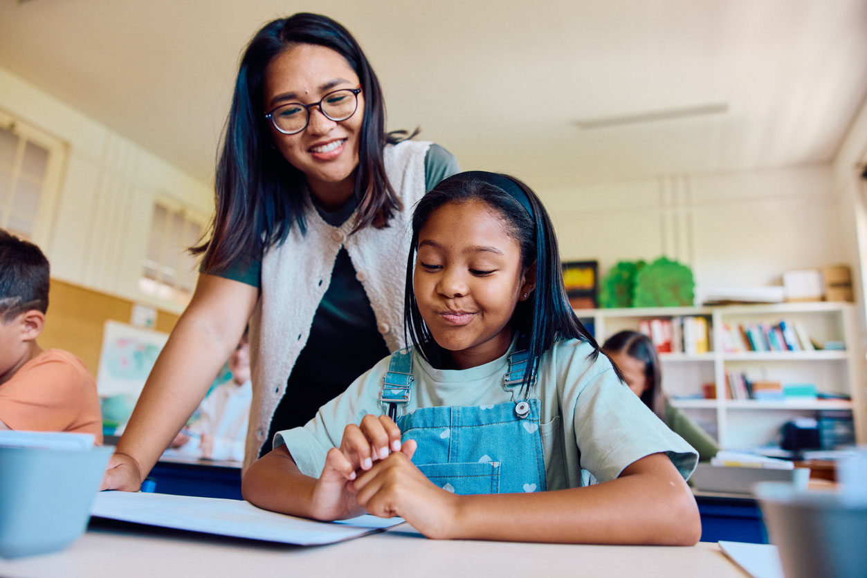 Helpful teacher giving attention to smirking schoolgirl at her desk during classroom lesson Helpful teacher leaning over desk giving attention to a smirking schoolgirl at her desk, assisting her complete a worksheet during classroom activity.