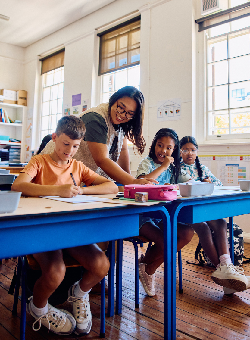 Full length shot of teacher assisting students writing in notebooks at desks in bright classroom